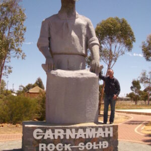 A bronze statue of a figure with a wide-brimmed hat and a man standing beside it under a clear sky, with the inscription "carnamah rock solid". Sculptor - Robert C Hitchcock