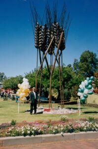 A man in a suit stands beside a tall bronze sculpture adorned with balloon bouquets, in an outdoor setting with clear skies. Sculptor - Robert C Hitchcock