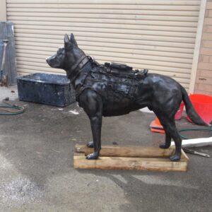 A black, life-sized bronze sculpture of a dog with a harness, positioned on a wooden platform in an outdoor setting with various cleaning tools nearby. Sculptor - Robert C Hitchcock